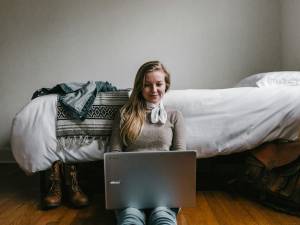 woman sitting beside a bed while using a laptop